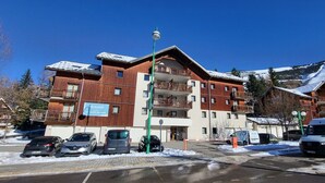 A multi-story building with balconies, a parking lot with several cars, and a snow-covered mountain in the background.