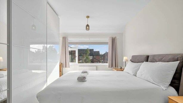 A modern hotel room with a large bed, a glass partition, a view of the outside, and a ceiling light.