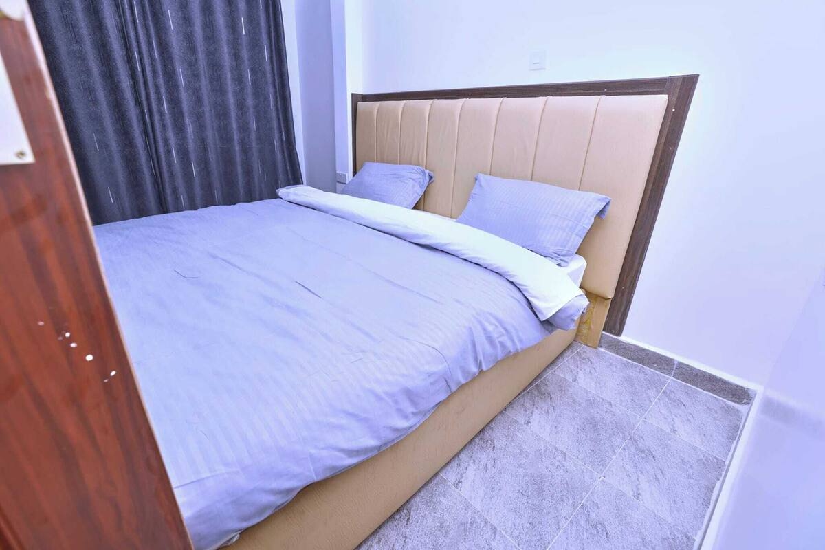 A neatly made bed with a headboard, a dark curtain partially visible, and a tiled floor.