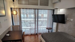 Et hotelværelse med en seng, et skrivebord med to fjernbetjeninger, en stol, et rundt sofabord, et fjernsyn ophængt på væggen og et vindue med let gardin.