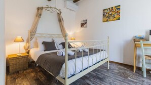 A bedroom with a canopy bed, a bedside table with a lamp, a wooden floor, and a desk with a chair. - Chambre d'hôtes - B&B - Fleur de Coton (Lagnes)