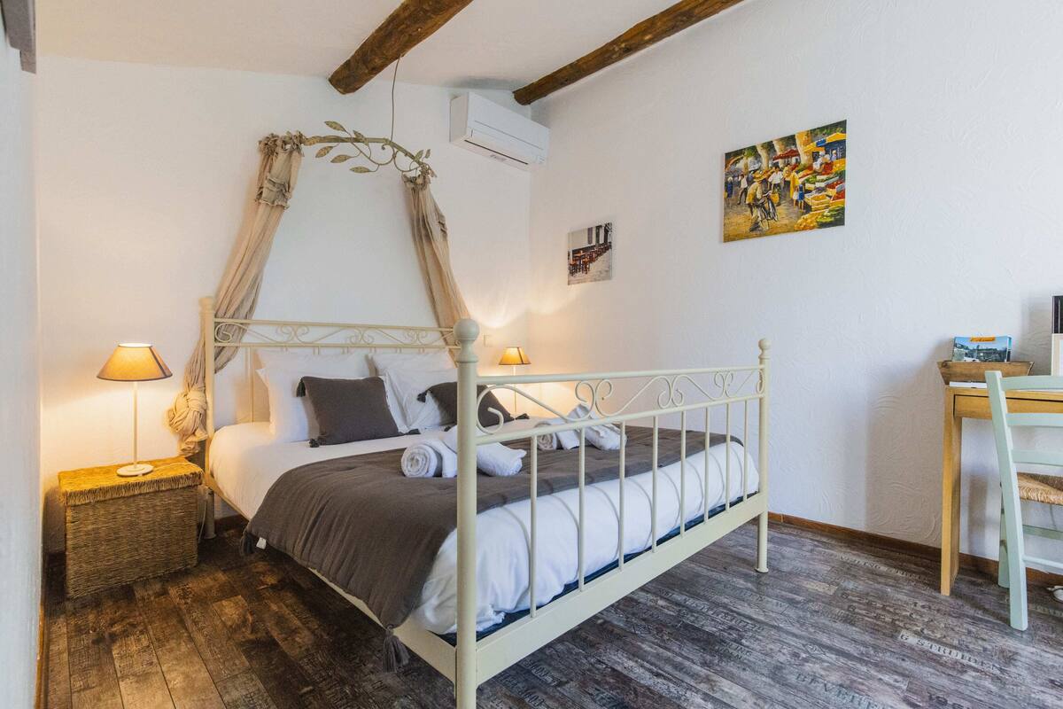 A bedroom with a canopy bed, a bedside table with a lamp, a wooden floor, and a desk with a chair.