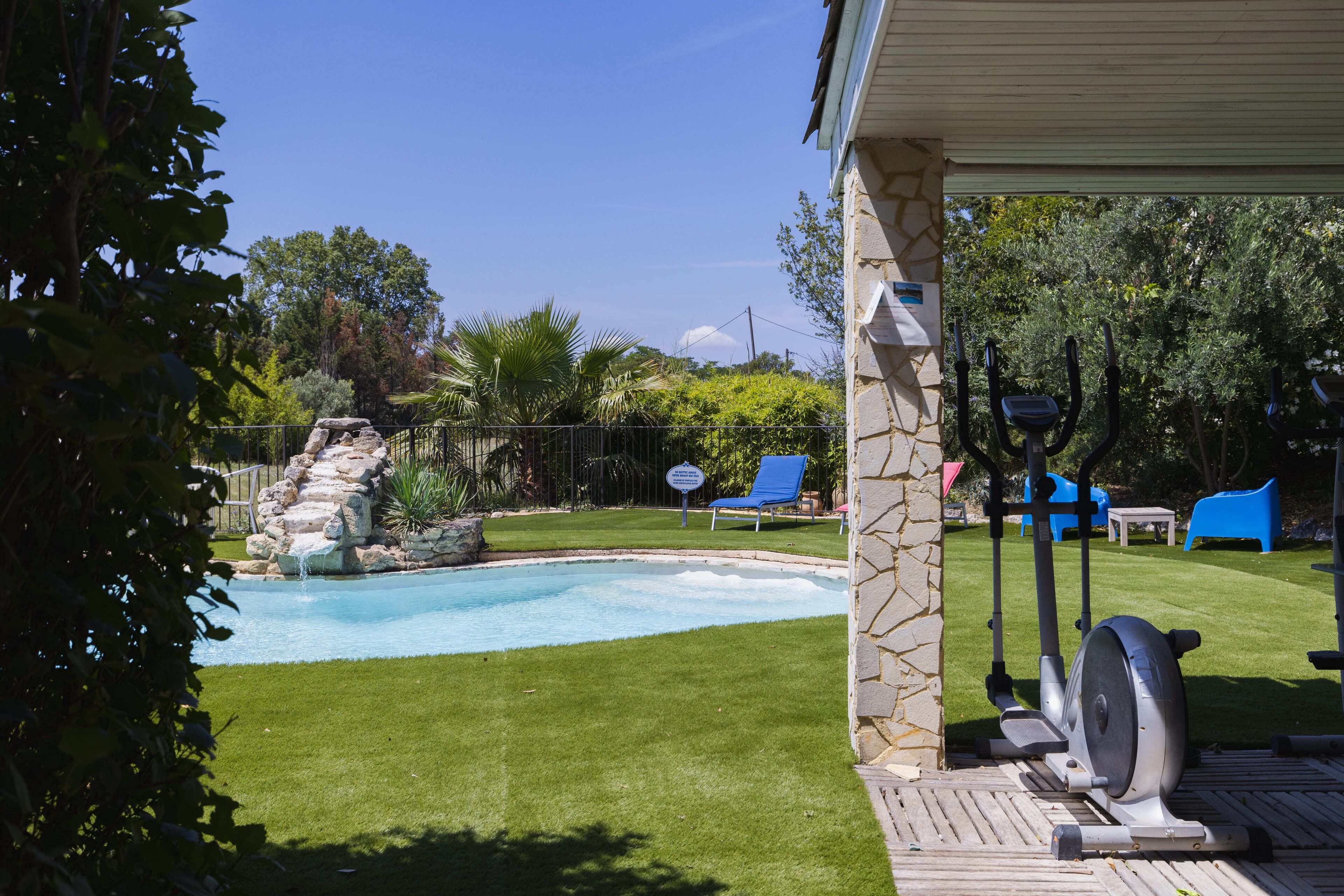 A backyard with a swimming pool, a patio area with exercise equipment, and a stone wall.