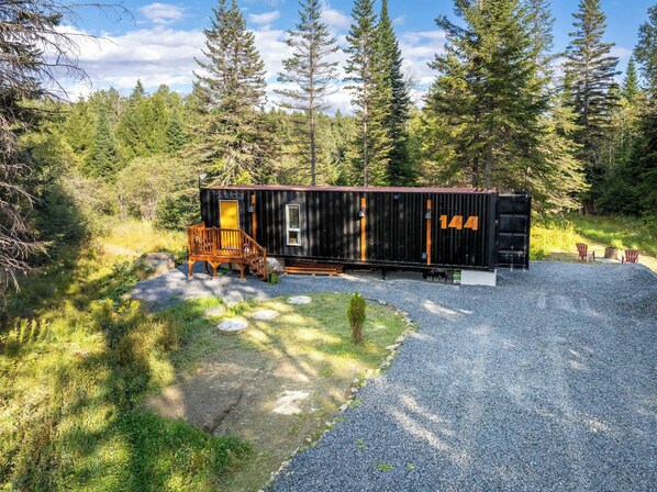 A rustic cabin with a wooden deck, a small porch, and a gravel driveway. - Spa & River View | Le Conteneur Caché (Saint-Magloire)