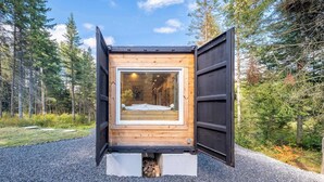 A small, open wooden cabin with a large window, surrounded by a forest.