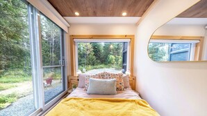 A bedroom with a large window, a bed with a yellow blanket, and a view of a garden.
