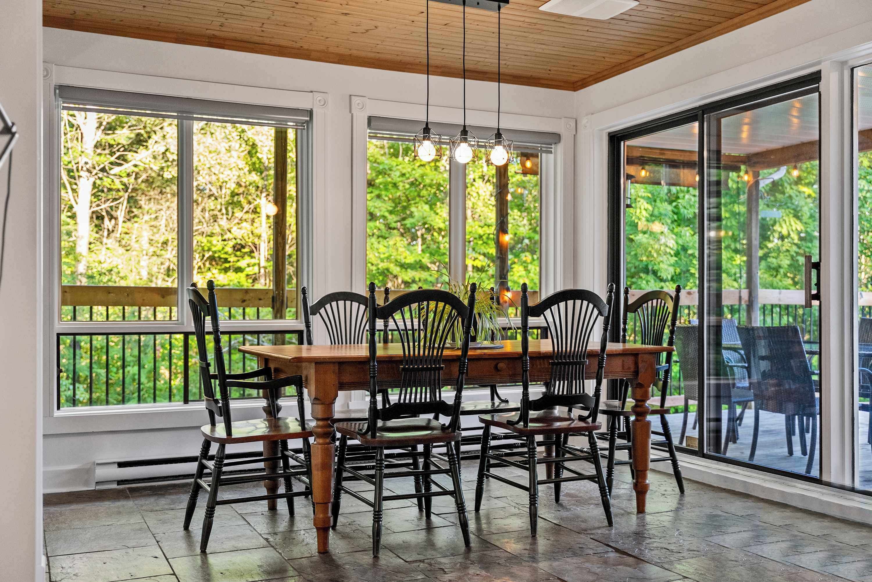A dining area with a wooden table and chairs, large windows, and a view of greenery.