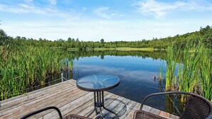 Una terraza de madera con una mesa de vidrio y sillas de mimbre, con vista a un lago tranquilo rodeado de altas hierbas y árboles.
