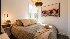 A bedroom with a bed, pillows, a bedside lamp, a plant, a hat, a framed picture, and a window.