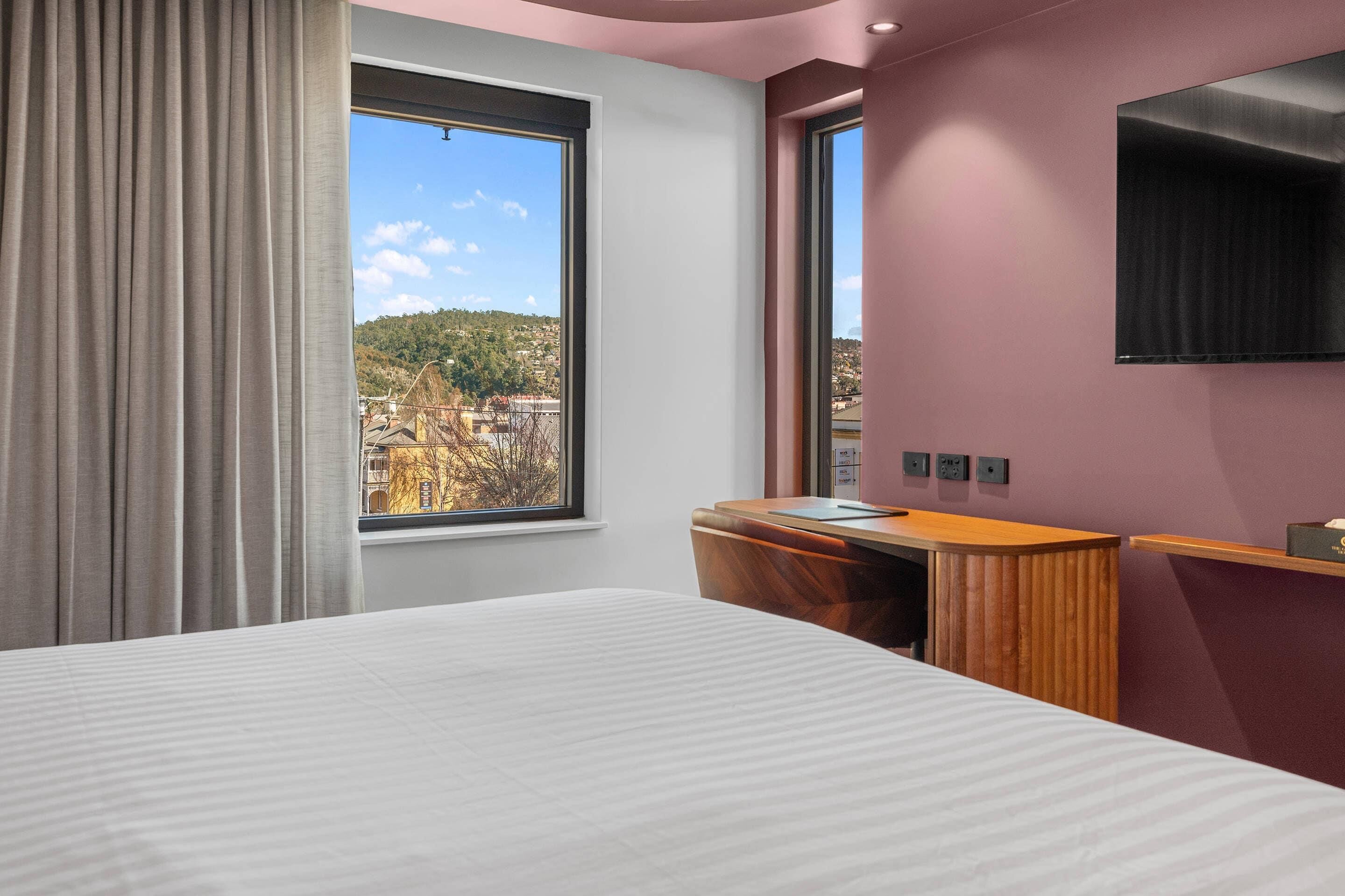 Une chambre d’hôtel équipée d’un lit, d’un bureau, d’une télévision, d’une fenêtre donnant sur des bâtiments et des collines, et d’une prise de courant fixée au mur.