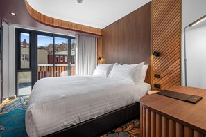 Een moderne hotelkamer met een groot bed, een houten bureau en uitzicht op de stad door een schuifglazen deur.