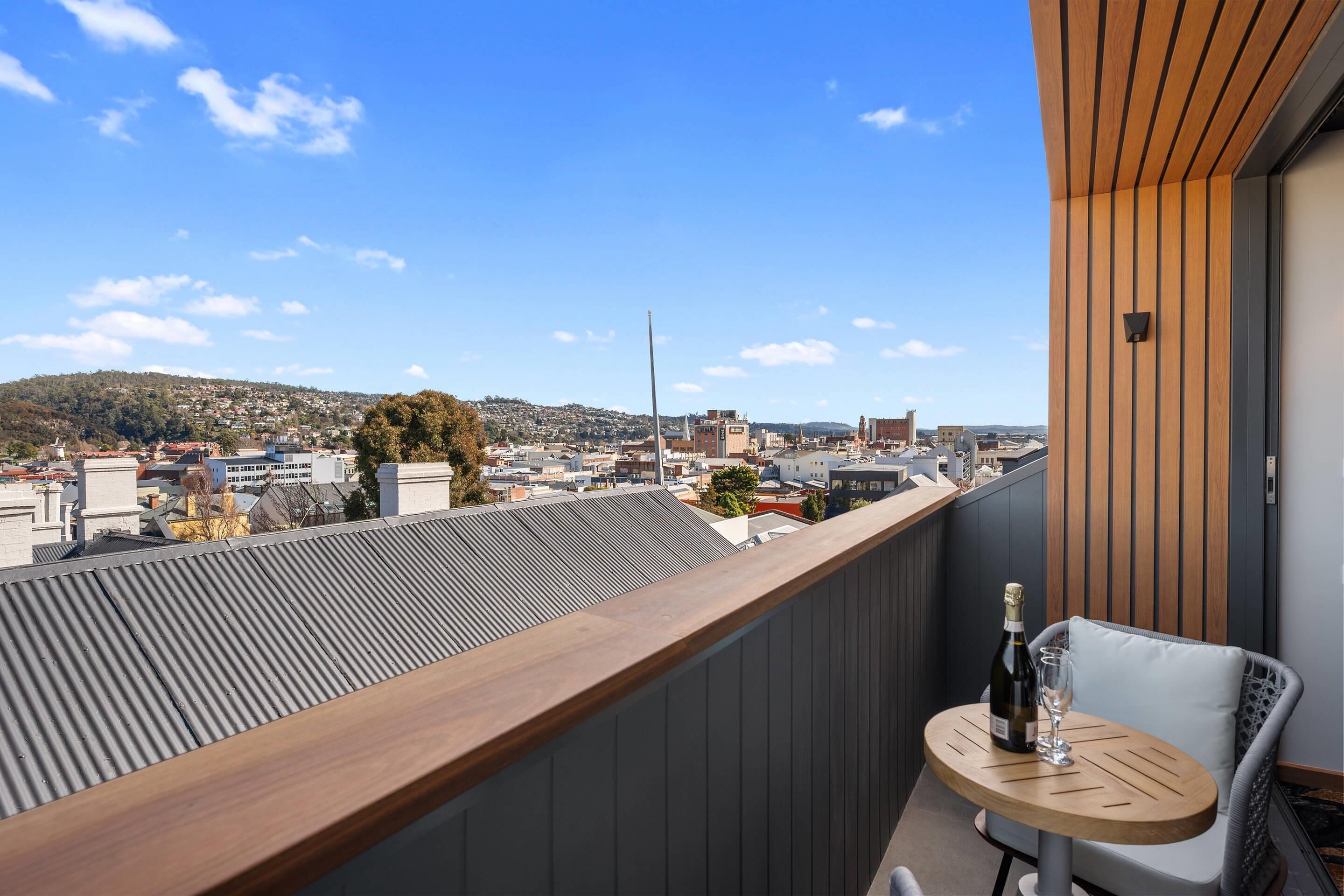 A balcony with a view of a cityscape, a wooden table with a bottle and glass, and a white chair.