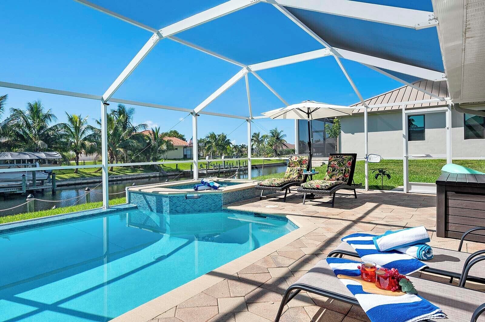 Ein Poolbereich mit überdachter Terrasse, Liegestühlen und Blick auf ein Gewässer und Grünflächen.