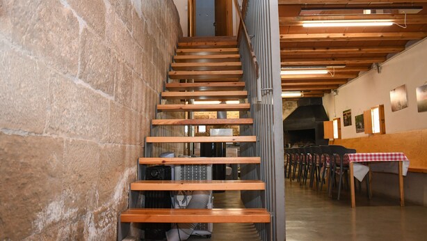 Una escalera de madera con un muro de piedra a un lado y una fila de sillas con mantel a cuadros al otro.