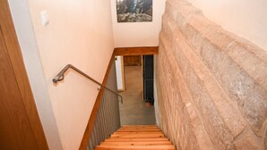 Una escalera de madera con barandilla de metal, que conduce a una habitación con pared de piedra y un cuadro en la pared.