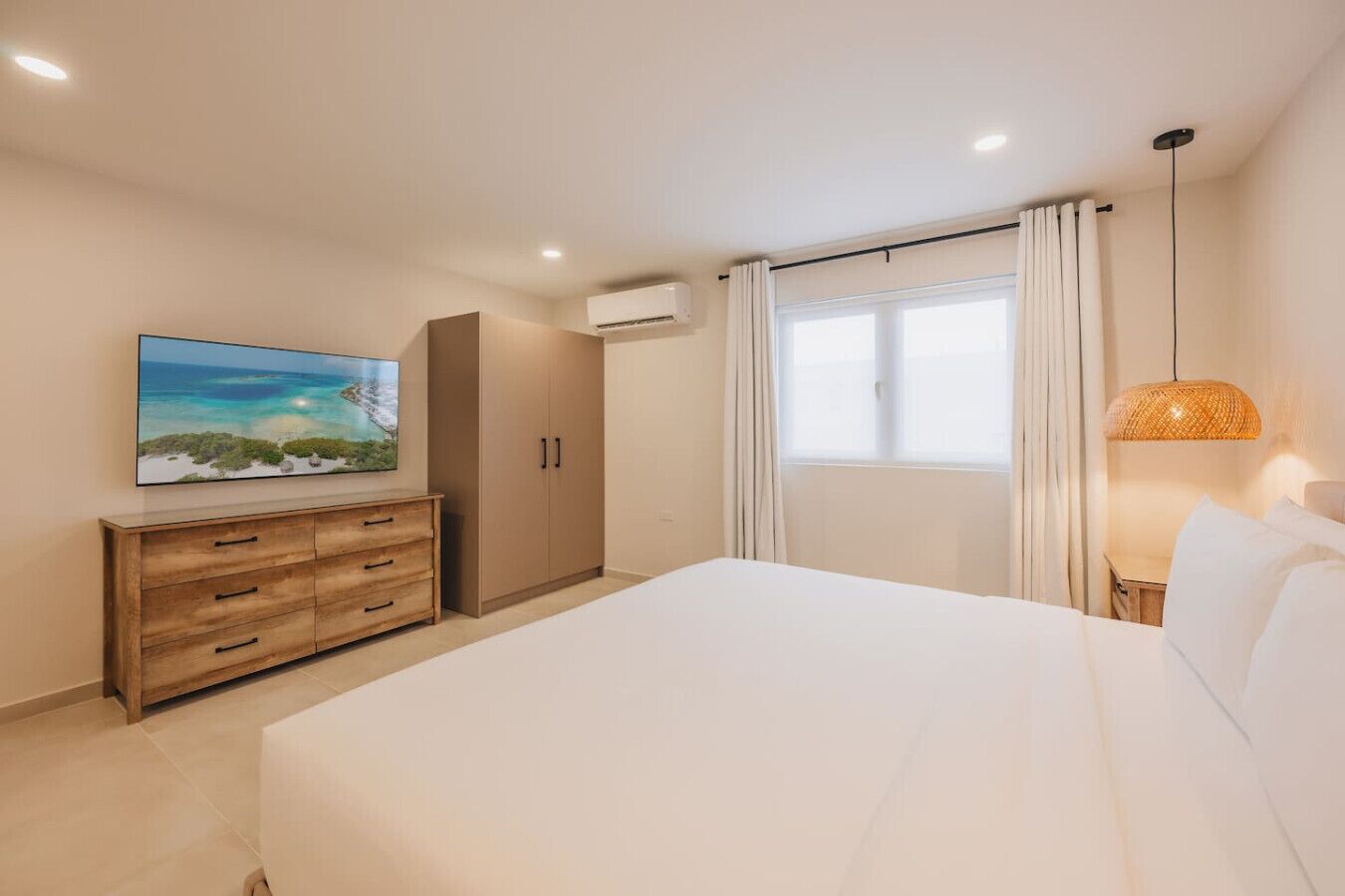 Une chambre à coucher comprenant un lit, une commode en bois, un tableau, un appareil de climatisation, une fenêtre avec des rideaux et une lampe suspendue.