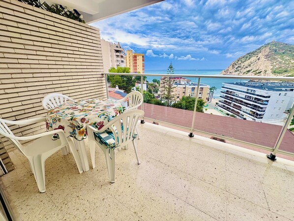 A balcony with a table and chairs, a tiled floor, and a view of buildings and a coastline.