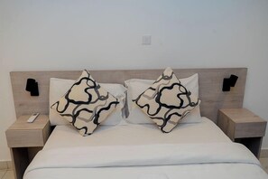 A bed with white bedding and two patterned pillows, flanked by nightstands with a remote control on one. - Loxwood House (Accra)
