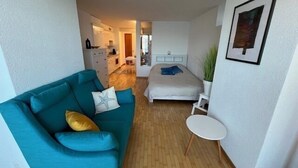 Ein kompakter Wohnbereich mit einem salbeifarbenen Sofa, einem kleinen runden Tisch, einem Bett und einer Küchenzeile im Hintergrund.