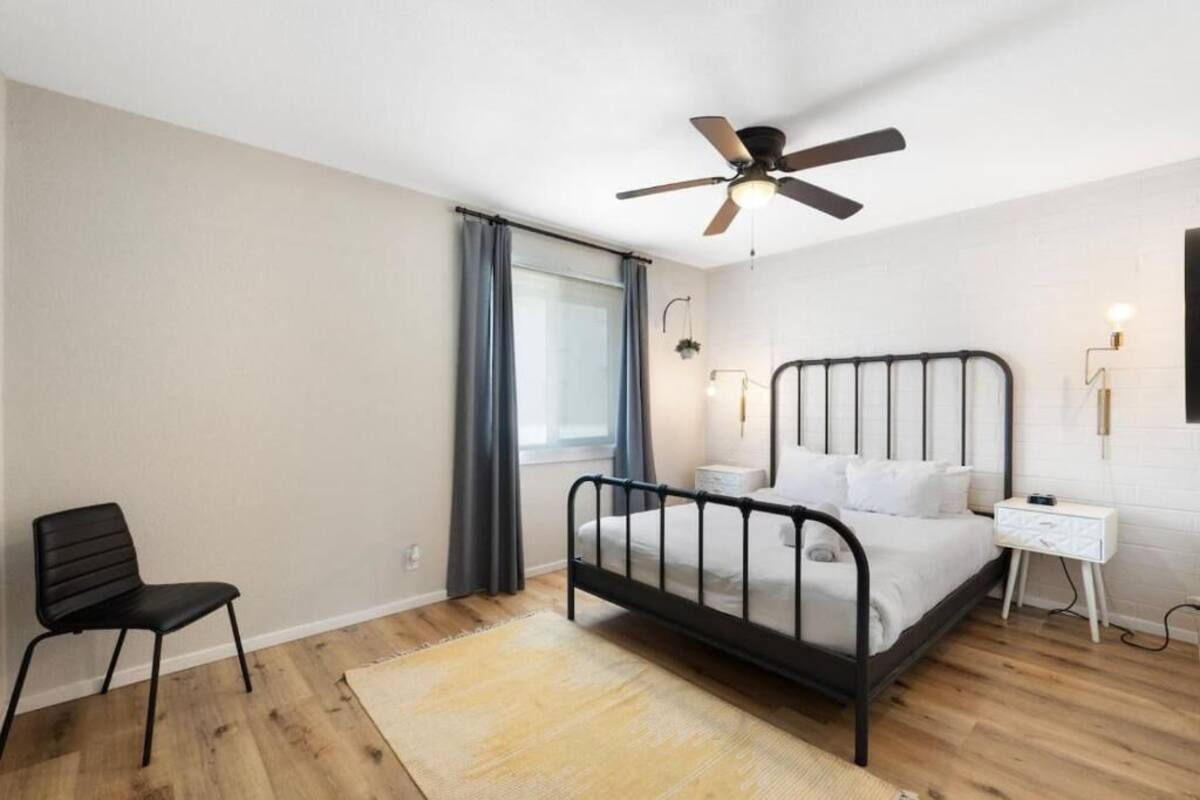 Un dormitorio con cama, una silla, ventilador de techo, ventana con cortinas y una mesita de noche.
