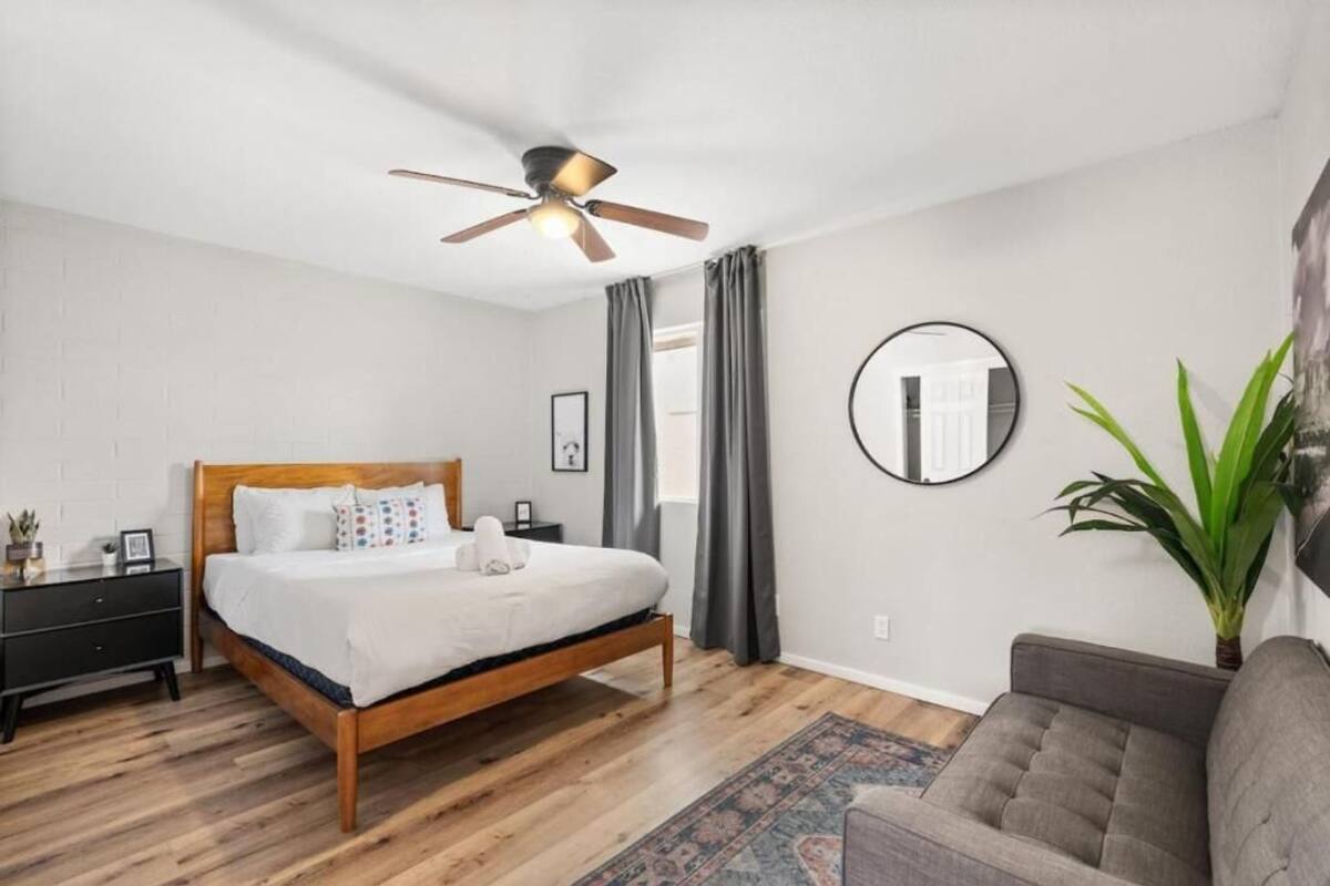 Un dormitorio con una cama de madera, un ventilador de techo, un espejo redondo, un sofá gris y una planta en maceta.