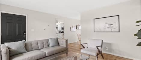 Ein Wohnzimmer mit Sofa, Sessel und Couchtisch. An der Wand hängt ein gerahmtes Bild, im Hintergrund ist eine Tür zu sehen.