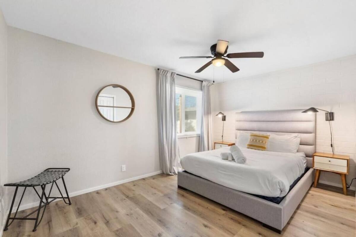 Un dormitorio con cama, ventilador de techo, mesita de noche, lámpara, ventana con cortinas y un taburete pequeño.