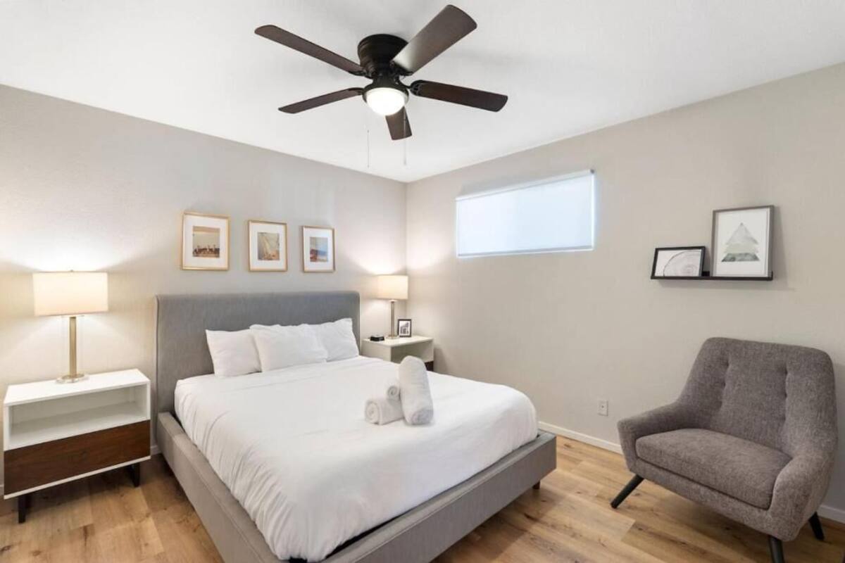 Un dormitorio ordenado con cama, mesitas de noche, una silla, ventilador de techo e imágenes enmarcadas en la pared.