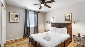 Ein Hotelzimmer mit einem hölzernen Bett, einem Deckenventilator, einem Nachttisch mit Lampe sowie zwei gerahmten Bildern an der Wand.