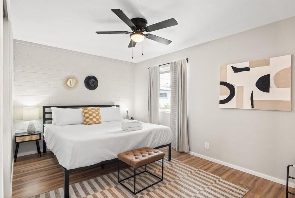 A bedroom with a bed, a ceiling fan, a bedside table, a bench, and a window with curtains.
