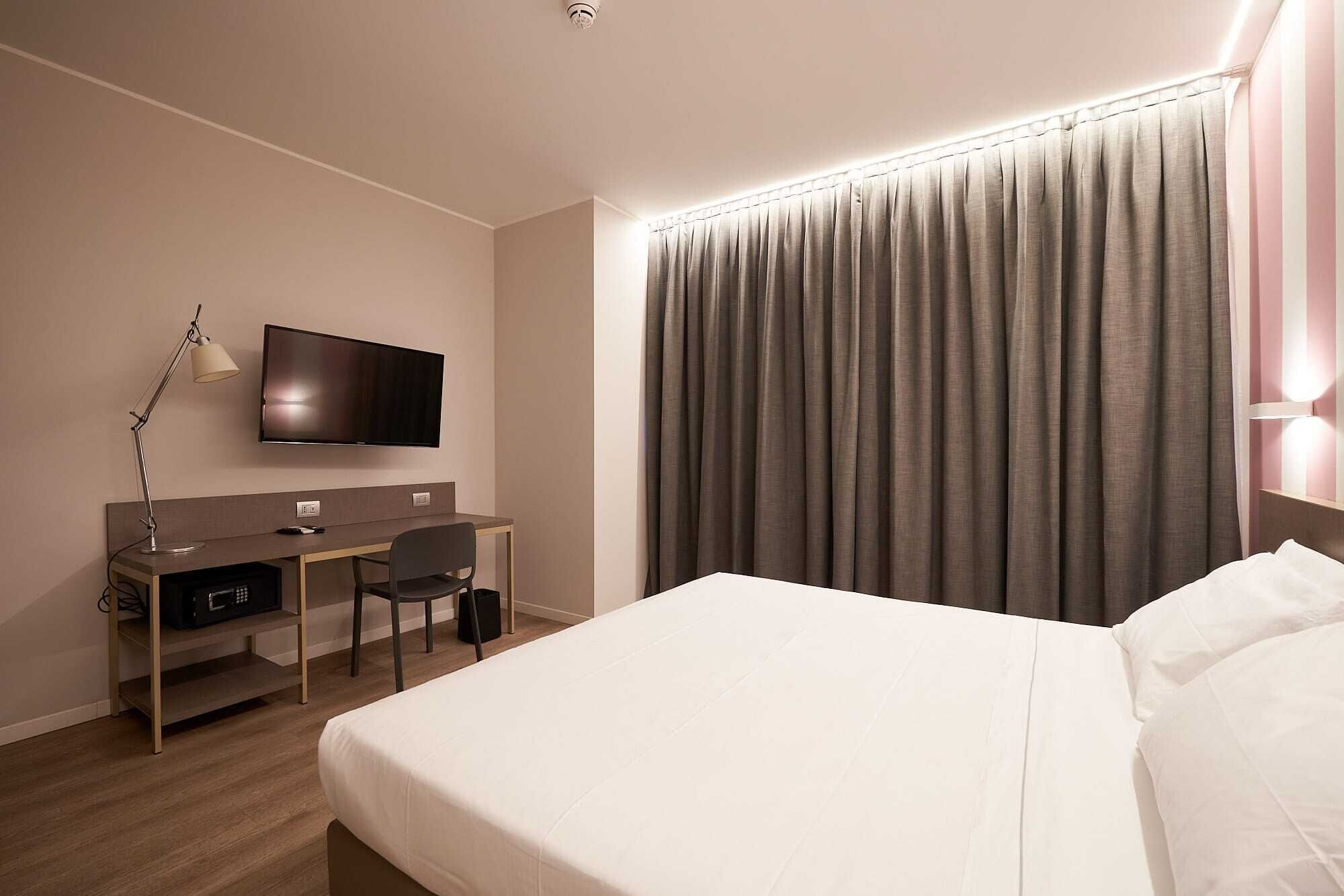 Una habitación de hotel con cama, escritorio con silla, televisor y un ventanal con cortinas.