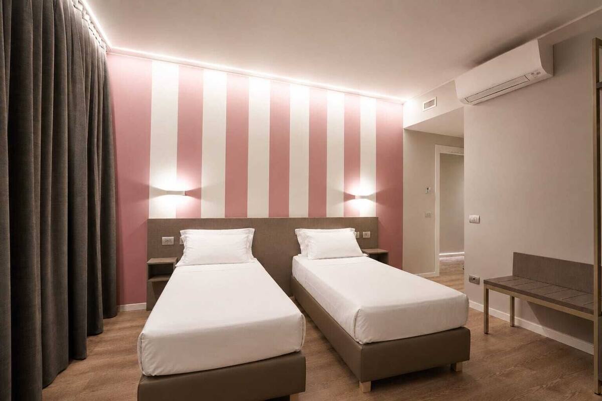 Habitación de hotel con dos camas, un banco, aire acondicionado en la pared y una puerta que da a otra habitación.