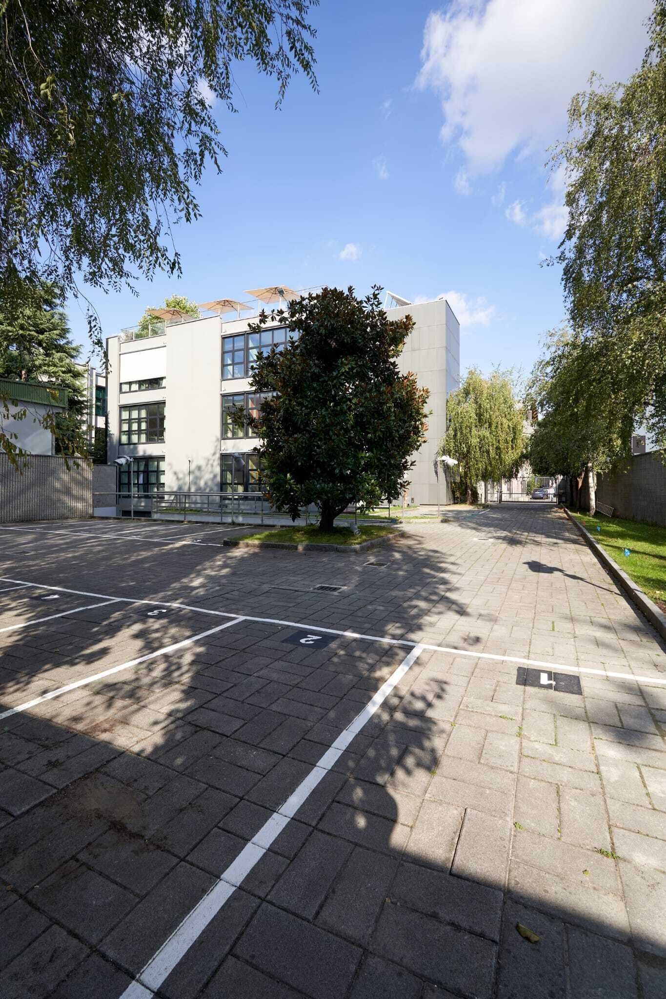 Ein Parkplatz mit eingezeichneten Stellplätzen, ein modernes Gebäude mit großen Fenstern und ein Baum im Vordergrund.