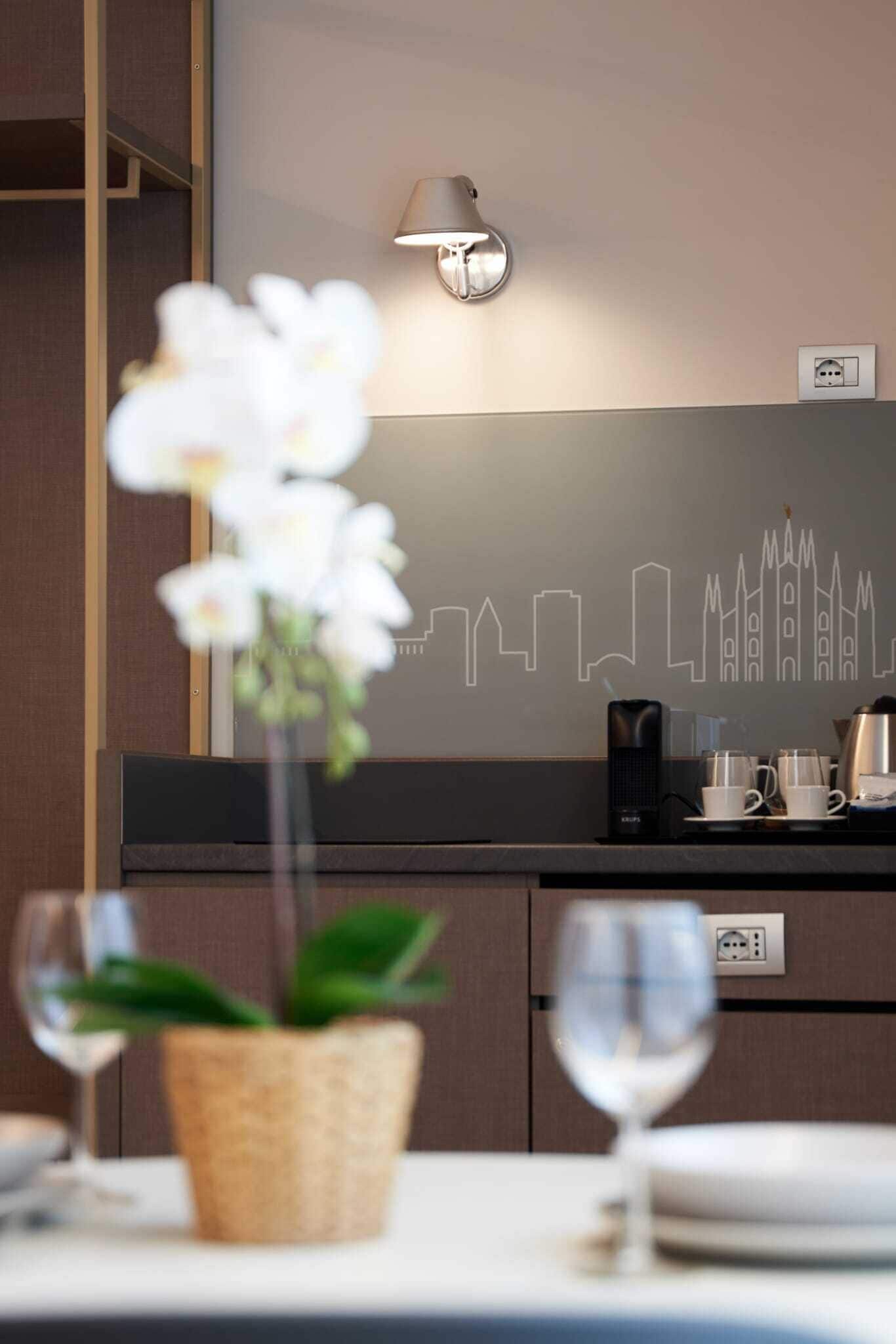 Una habitación de hotel con un cuadro de un paisaje urbano, una mesita de noche con una planta, una cafetera y una lámpara.