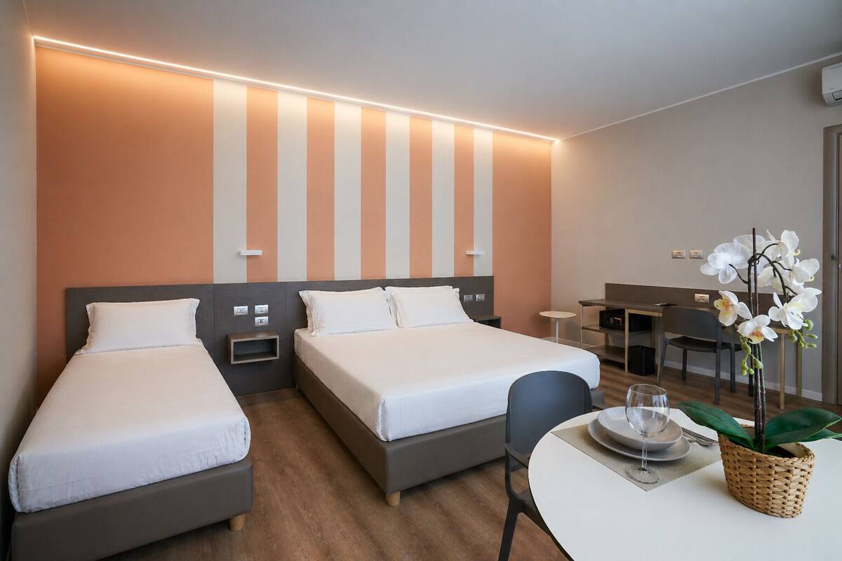 Una habitación de hotel con dos camas individuales, un escritorio pequeño, una silla, una mesa de comedor con un jarrón de flores y un aire acondicionado instalado en la pared.