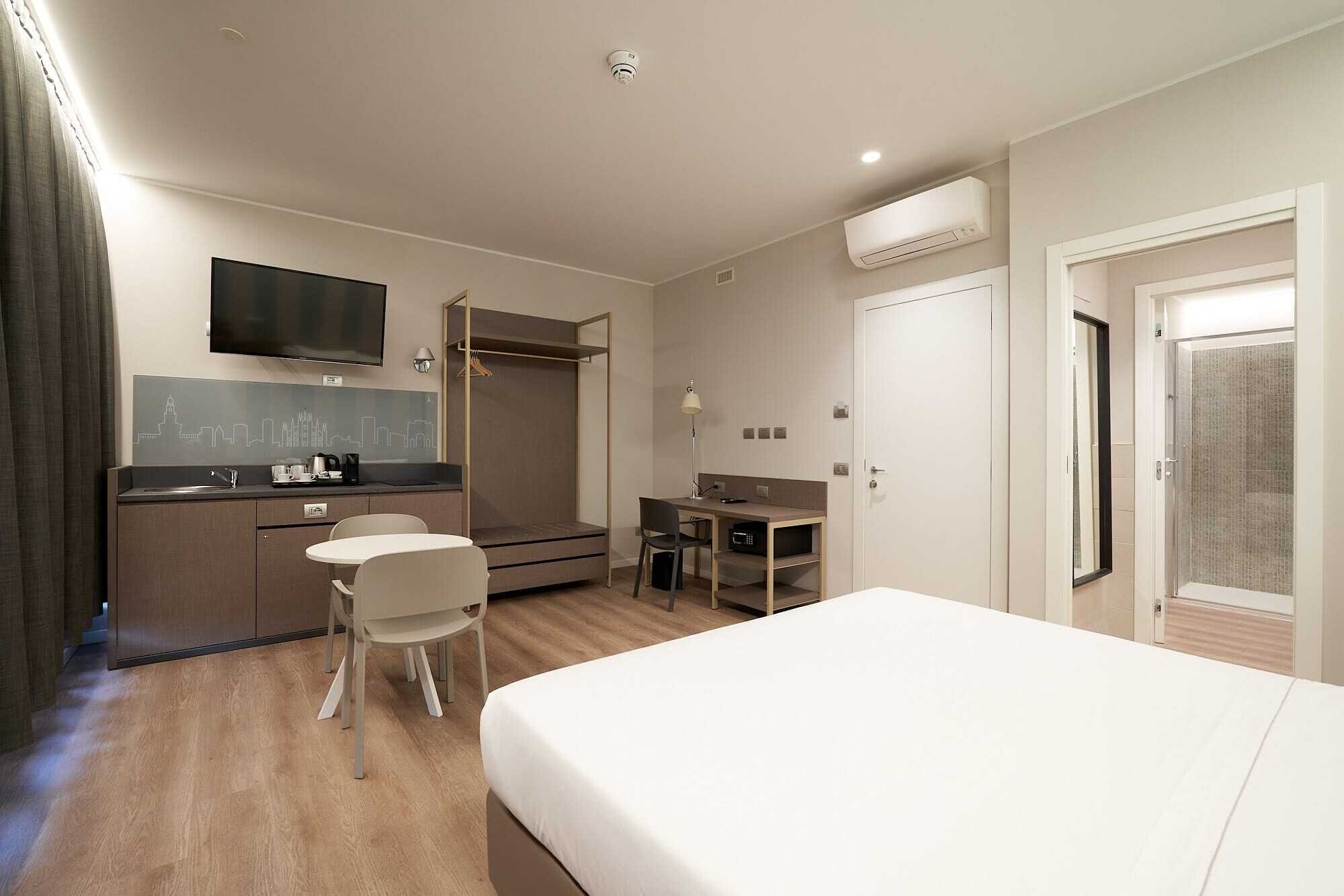 Ein Hotelzimmer mit Bett, Schreibtisch, Stuhl, einer kleinen Küchenzeile und einem Fernseher.