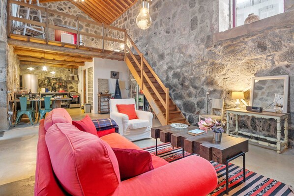 A spacious living area with a red sofa, wooden staircase, stone walls, and a kitchen visible in the background.