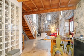 A rustic stone interior with a wooden staircase, a dining area with chairs, a table, and a wine bottle.