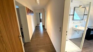 A hallway with a wooden door on the left, a glass door leading to a bathroom with a sink and mirror, and a tiled floor. - Tanne 16 (Braunlage)