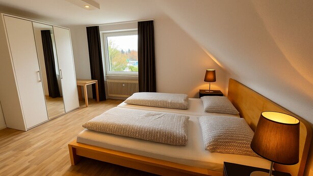 A modern bedroom with a large bed, bedside lamps, a mirror, and a view of trees outside. - Tanne 16 (Braunlage)