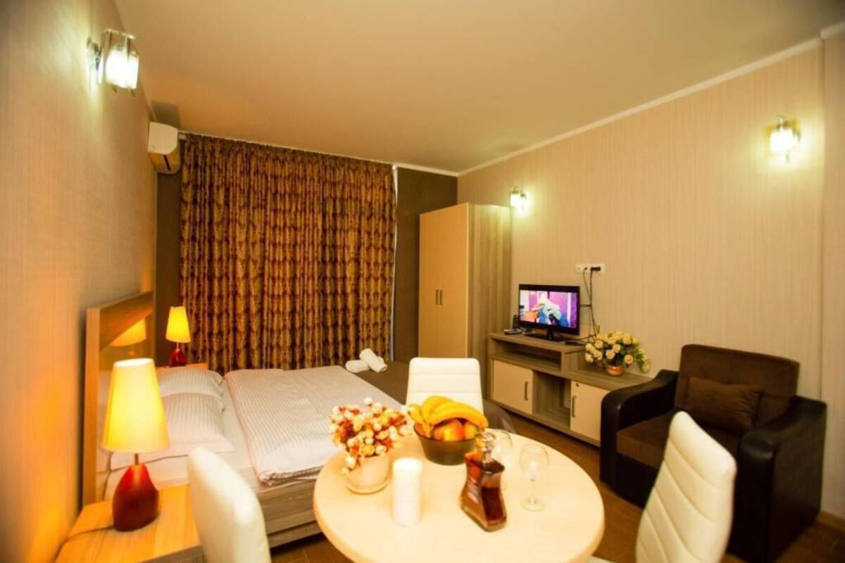 A hotel room with a bed, bedside table with a lamp, a small round table with a bowl of fruit, a bottle, and a glass. There is a TV on a stand, a chair, and a cabinet with a mirror.