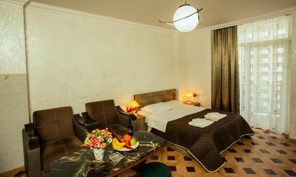 A hotel room with a bed, two armchairs, a small table with fruit and flowers, a nightstand with a lamp, and a window with curtains.