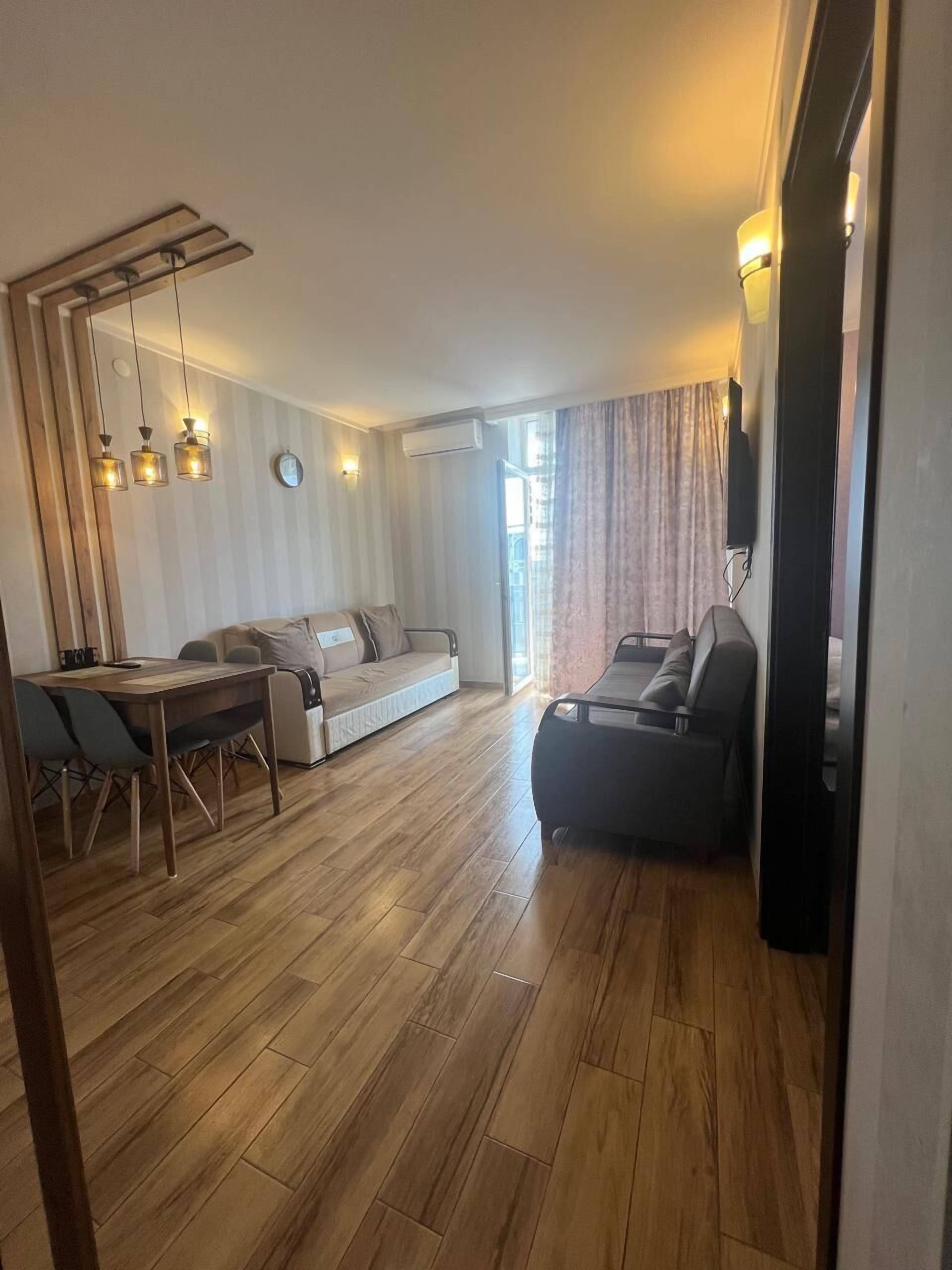 A modern living room with a wooden floor, a sofa, a dining table with chairs, and a TV mounted on the wall.