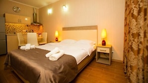 A hotel room with a bed, two bedside tables with lamps, a small desk with a chair, a kitchen area, and a patterned curtain. - Batumi Royal Apartments (Batumi)