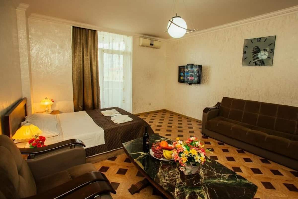 A hotel room with a bed, a sofa, a dining table with fruit and flowers, a lamp, and a clock on the wall.