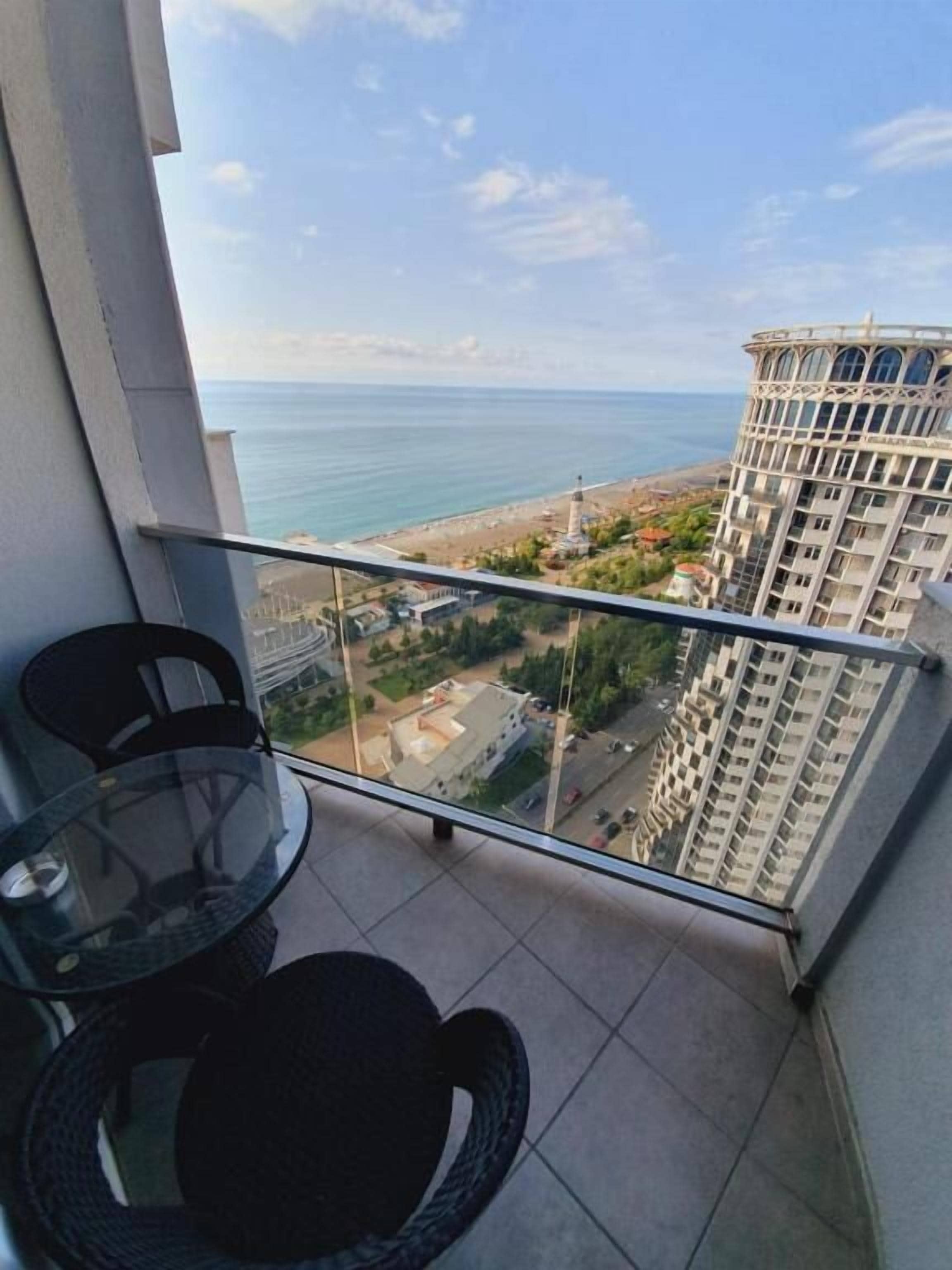 Un balcón con una mesa y sillas de vidrio, con vistas a la playa y un edificio alto.
