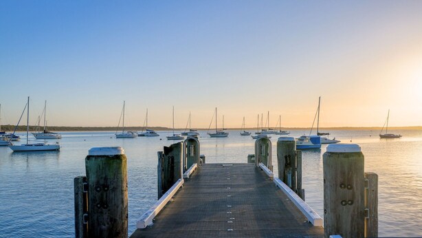 A wooden pier leading to a calm harbor with several sailboats anchored in the distance. - Coastal Drift – your Jervis Bay escape starts here (Callala Bay)