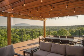 Una terraza exterior con piso de madera, una pérgola con luces colgantes y vistas a un paisaje boscoso.