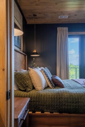 Una cama con una manta verde, varios almohadones, una lámpara colgante, una ventana con cortinas y vistas al exterior con vegetación.