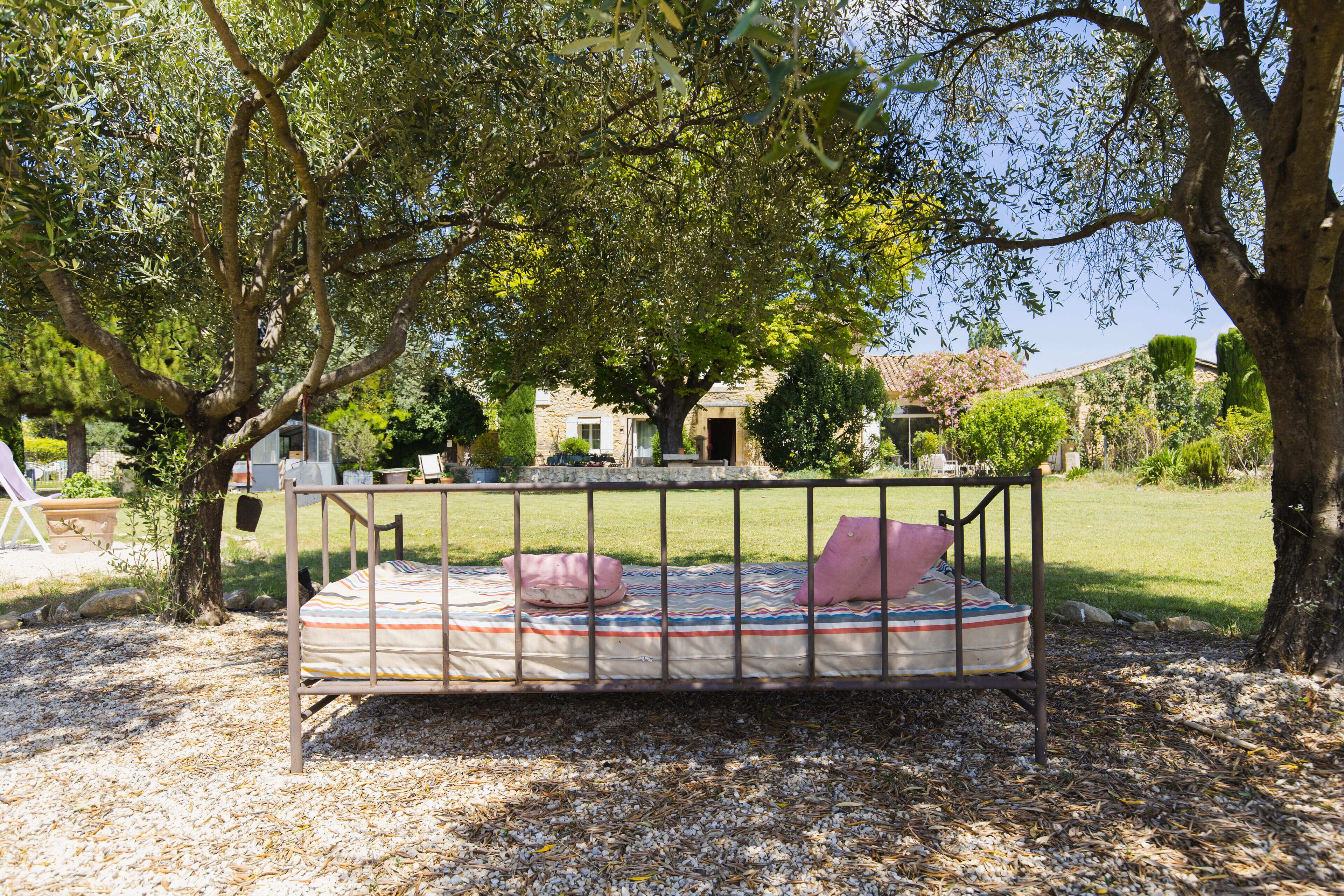 Una cama con estructura metálica y colchón a rayas, rodeada por una zona de grava y árboles.